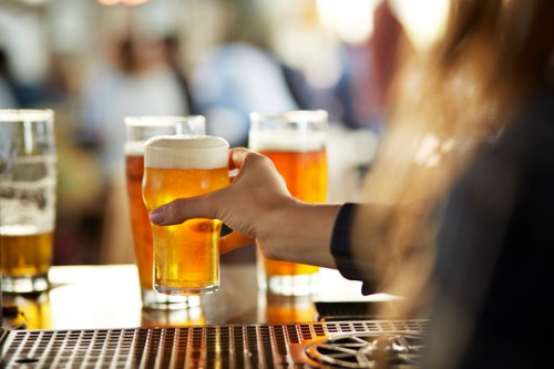 A hand holding a glass of beer, with several other glasses in the background on a bar counter.