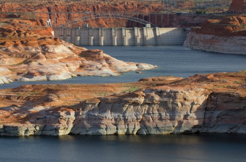A dam spans a reservoir surrounded by rocky, arid terrain under a clear blue sky.