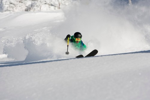 A skier in a green jacket and black helmet carves through fresh powder snow, creating a spray of snow around them.
