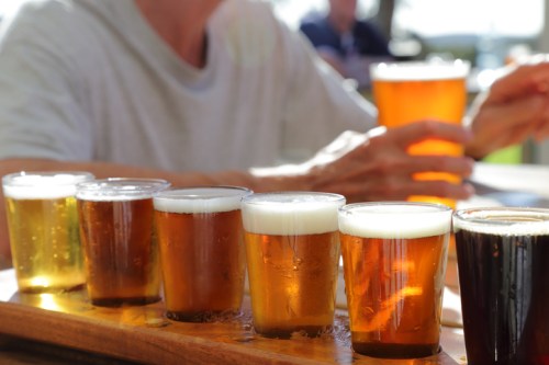 A flight of various beers in small glasses on a wooden tray, with a person holding a pint in the background.
