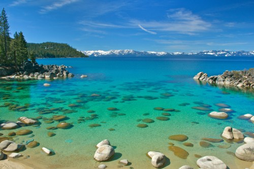 A serene lake with clear turquoise water, rocky shores, and snow-capped mountains in the background under a blue sky.