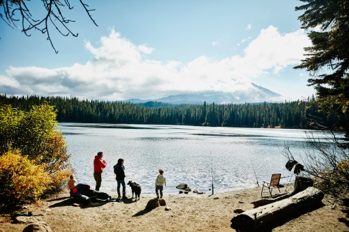 A family by a serene lake, with a dog, surrounded by trees and mountains under a clear blue sky.