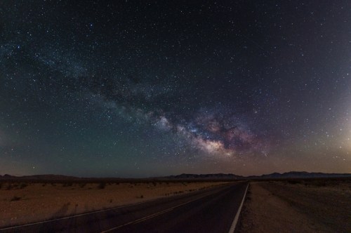 A starry night sky with the Milky Way arching over a deserted road in a desert landscape.