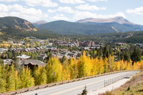A scenic view of a mountain town surrounded by autumn foliage and distant peaks under a partly cloudy sky.