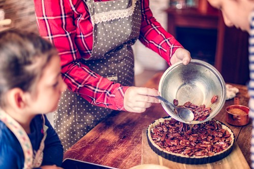 A person in an apron adds pecans to a pie crust while a child watches attentively in a cozy kitchen setting.