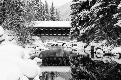 A snow-covered landscape featuring a bridge over a calm river, surrounded by trees and rocky banks.