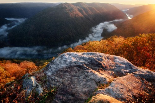 A scenic view of a valley at sunrise, with mist rising from a river and colorful autumn foliage on the hills.