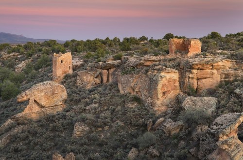 Ancient stone ruins atop rocky terrain, surrounded by shrubs and a colorful sunset sky.