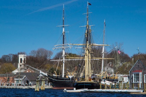 Two tall ships docked in a harbor, with a clear blue sky and historic buildings in the background.
