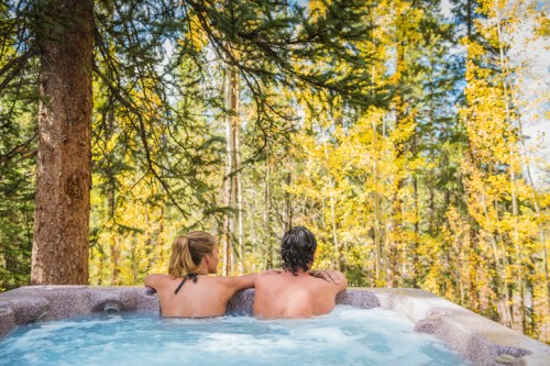 A couple relaxes in a hot tub surrounded by trees with autumn foliage.