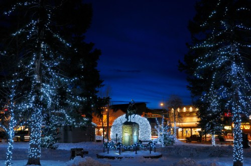 A winter scene featuring a statue surrounded by trees adorned with twinkling lights against a twilight sky.