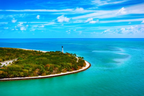 A scenic view of a coastal landscape with a lighthouse, lush greenery, and vibrant blue waters under a partly cloudy sky.