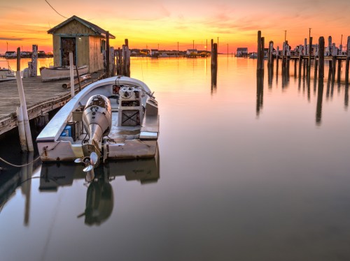 A calm sunset over a marina, featuring a boat docked by a wooden pier and reflections on the water.