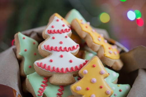 A close-up of festive Christmas tree-shaped cookies decorated with colorful icing, set against a blurred background of lights.