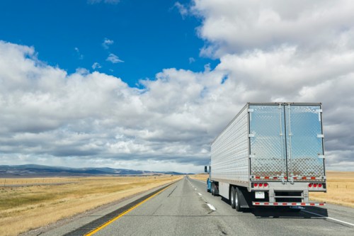 A large truck driving on a wide, empty highway under a cloudy blue sky.
