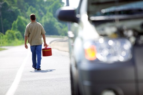 A person walks along a road carrying a red gas can, with a vehicle blurred in the foreground.