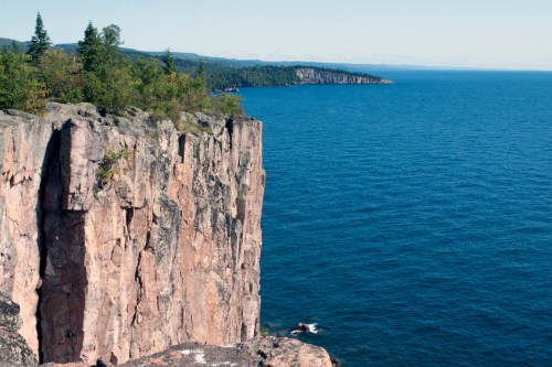 A rocky cliff overlooks a calm blue lake, surrounded by trees and distant hills under a clear sky.