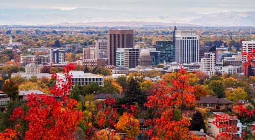 A panoramic view of a city skyline surrounded by autumn foliage, showcasing colorful trees and distant mountains.