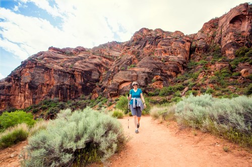 A hiker walks along a dirt trail surrounded by rocky cliffs and green shrubs under a partly cloudy sky.