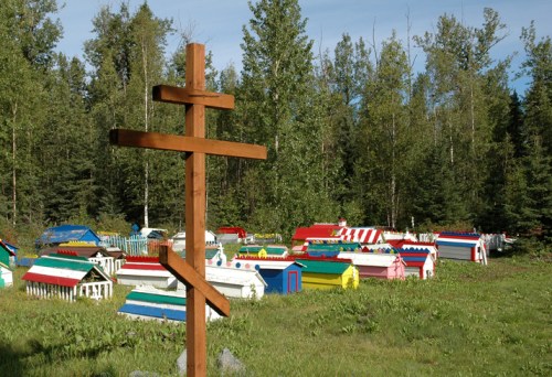 A wooden cross stands in front of colorful, small houses in a forested area, surrounded by trees and greenery.