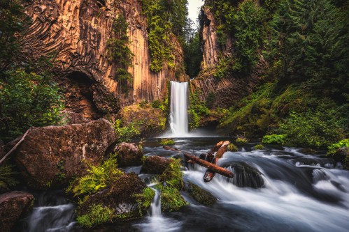 A serene waterfall cascades into a river, surrounded by lush greenery and rocky cliffs.
