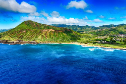 Aerial view of a lush green mountain overlooking a blue ocean and sandy beach under a bright blue sky.