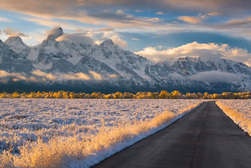 A winding road leads through a snowy landscape with majestic mountains and golden trees under a dramatic sky.