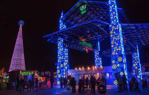 A festive scene with colorful holiday lights, a tall illuminated tree, and people gathered near a gift shop.