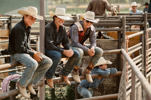Four cowboys in hats and boots sit on a fence, smiling and chatting at a rodeo event.