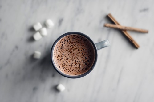 A top-down view of a mug filled with frothy coffee, surrounded by sugar cubes and cinnamon sticks on a marble surface.