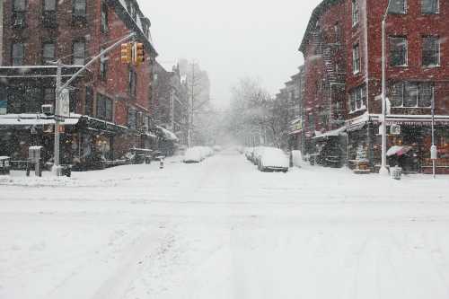 A snowy street scene with red brick buildings, covered cars, and heavy snowfall, creating a quiet winter atmosphere.