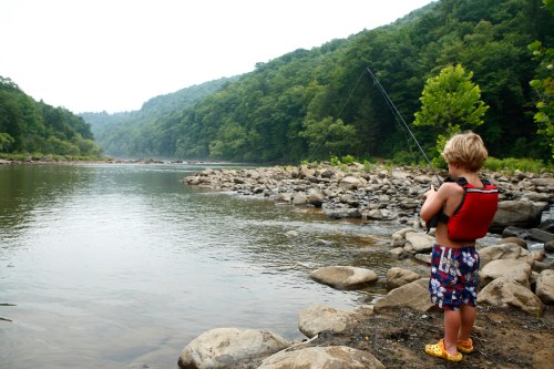 A child in a life jacket fishes by a river, surrounded by rocky shores and lush green hills.
