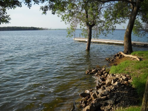A serene lakeside scene with trees, a wooden dock, and rocky shoreline under a clear sky.