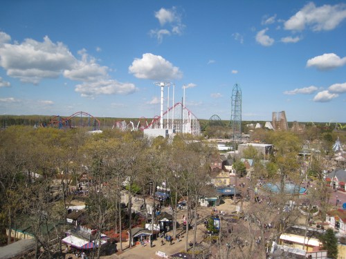 Aerial view of an amusement park with roller coasters, trees, and a clear blue sky with fluffy clouds.