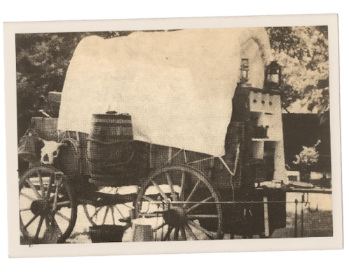A vintage wagon covered with a white tarp, featuring barrels and lanterns, set in a natural outdoor environment.
