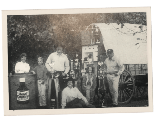 A black-and-white photo of a group of people with trophies, standing by a wagon and a large bottle of Head Country sauce.