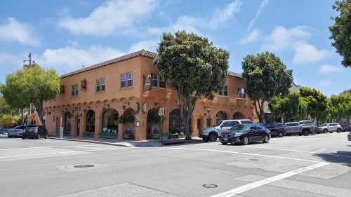 A charming, two-story building with a stucco facade and trees, located at a street intersection on a sunny day.