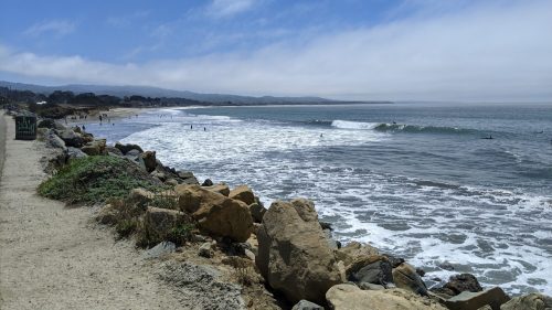 A scenic beach view with waves crashing, rocky shoreline, and surfers in the water under a blue sky with clouds.
