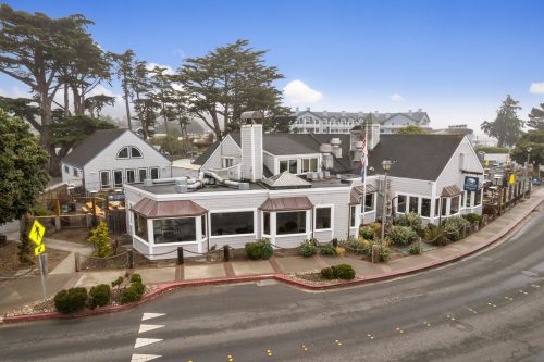 A coastal restaurant with a white exterior, surrounded by trees, on a street with a clear blue sky in the background.