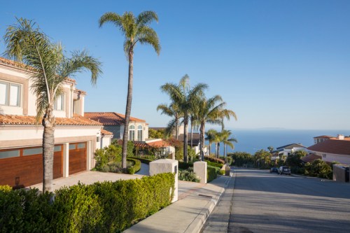 A scenic view of a residential street lined with palm trees, leading down to the ocean under a clear blue sky.