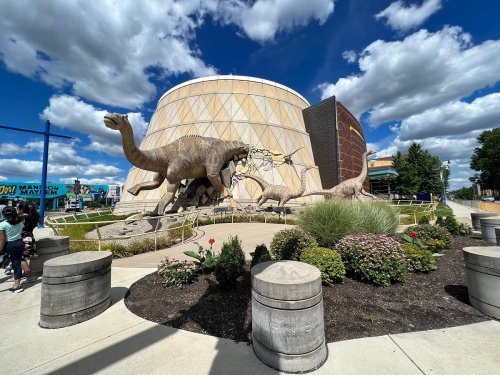 A large dinosaur-themed building with dinosaur sculptures outside, surrounded by flowers and a blue sky with clouds.