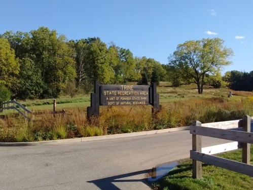 Sign for Trine State Recreation Area, surrounded by greenery and trees, with a clear blue sky above.