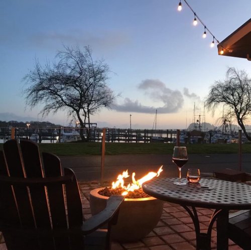 A cozy outdoor scene with a fire pit, wine glasses, and a view of a marina at sunset, illuminated by string lights.