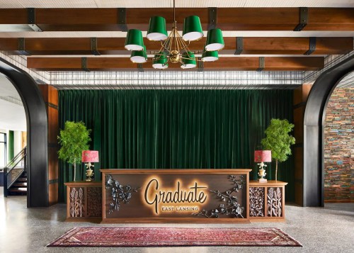 A stylish hotel lobby with a wooden reception desk, green curtains, plants, and decorative lighting.