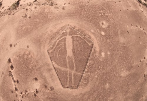 Aerial view of a large geoglyph resembling a human figure, surrounded by dry, barren land and faint circular patterns.