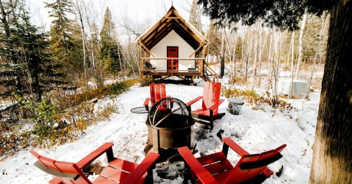 A cozy cabin in a snowy forest, surrounded by red chairs and a fire pit, with trees in the background.
