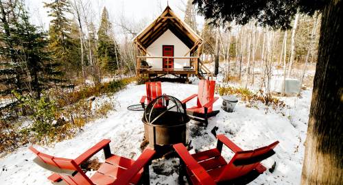 A cozy cabin surrounded by snow, with red chairs arranged around a fire pit in a forested area.