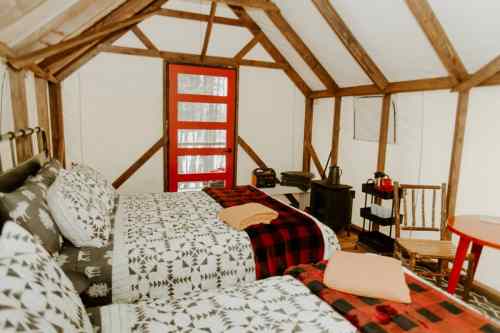 Cozy cabin interior with two beds, red door, wooden furniture, and a small stove, surrounded by nature.