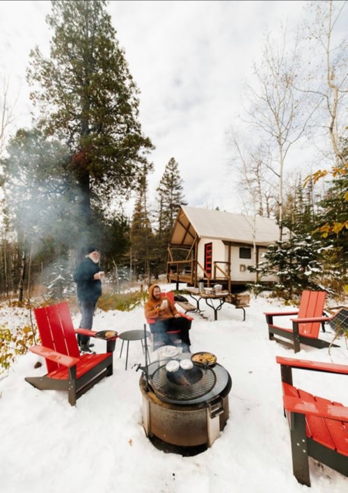 A cozy winter scene with two people by a fire pit, surrounded by snow and red chairs, near a cabin in the woods.
