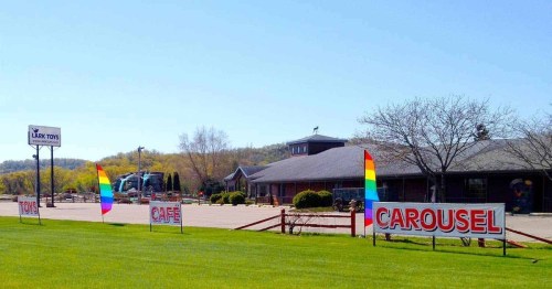 A colorful storefront with signs for "Toys," "Cafe," and "Carousel," surrounded by green grass and trees under a clear blue sky.